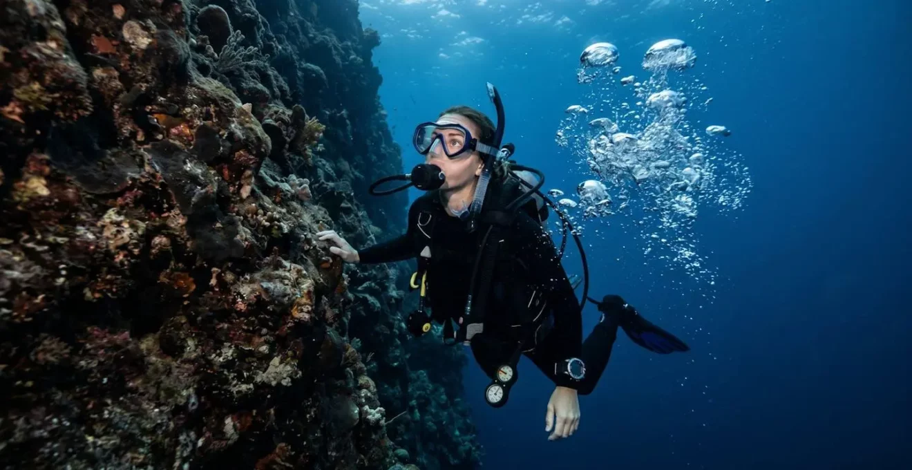 Plongeur en remontée lente le long d'une falaise sous-marine, expression de concentration visible à travers le masque, bulles irrégulières et rapprochées, fond bleu profond