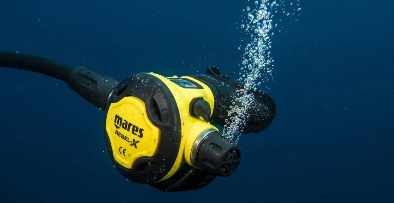 Gros plan sur un second étage de détendeur jaune et noir immergé, partiellement ouvert, filet de bulles s'échappant, fond bleu homogène